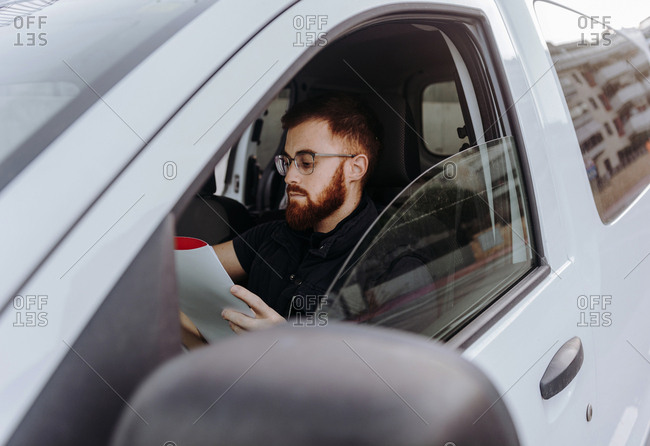 Side view of attentive thoughtful adult man focusing and checking documents while sitting behind steering wheel in car cabin during daytime on blurred background