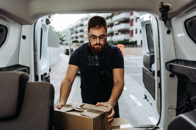 Attentive adult bearded courier in glasses carefully unloading cardboard boxes from car for further delivery to customer on blurred background during daytime