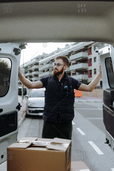 Attentive adult bearded courier in glasses carefully unloading cardboard boxes from car for further delivery to customer on blurred background during daytime