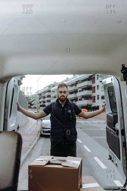 Attentive adult bearded courier in glasses carefully unloading cardboard boxes from car for further delivery to customer on blurred background during daytime