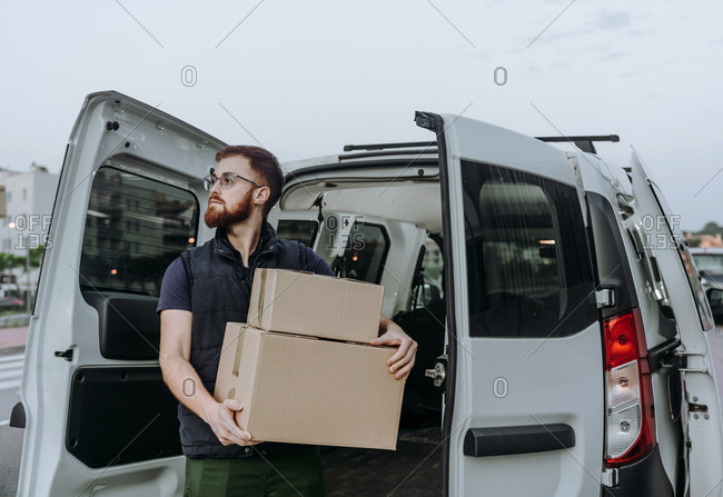 Attentive adult bearded courier in glasses carefully unloading cardboard boxes from car for further delivery to customer on blurred background during daytime