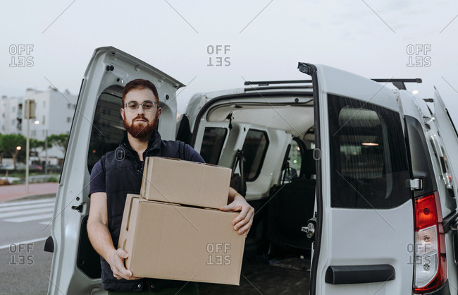 Attentive adult bearded courier in glasses carefully unloading cardboard boxes from car for further delivery to customer on blurred background during daytime