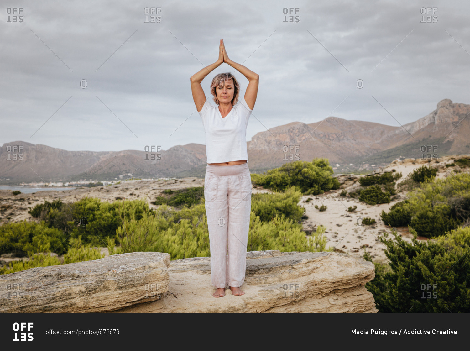 Full body barefoot adult female clasping hands over head and closing eyes while standing on rock in nature and meditating