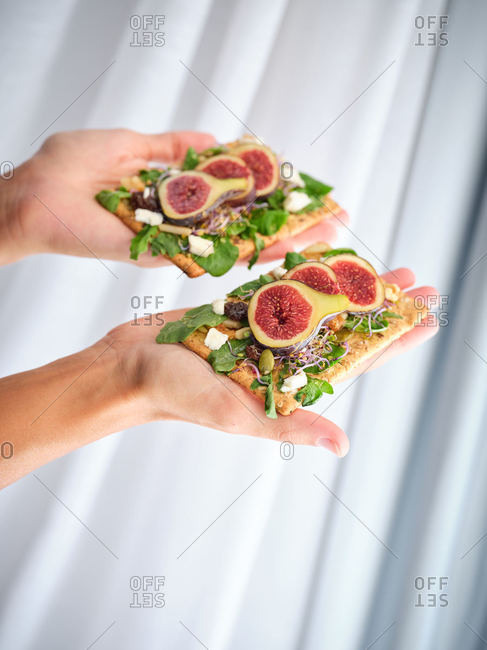 Crop person holding homemade colorful open sandwich with slices of fig and pieces of cheese on crisp rye bread with aromatic green leaves of rocket salad against white wall