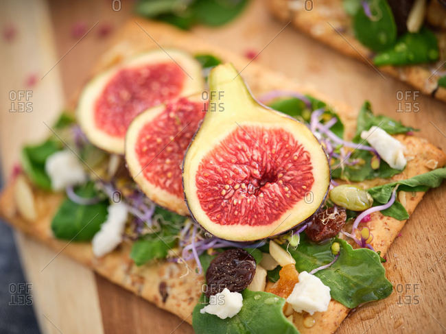 Ripe whole half and sliced figs among flavored green leaves of herbs and colorful flowers on wooden cutting board against blurred white wall
