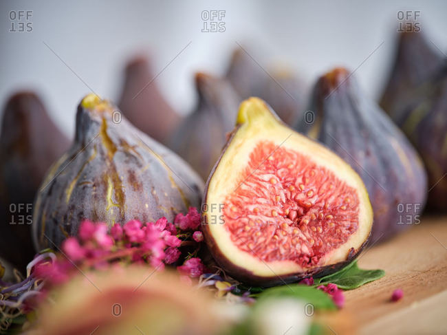 Ripe whole half and sliced figs among flavored green leaves of herbs and colorful flowers on wooden cutting board against blurred white wall