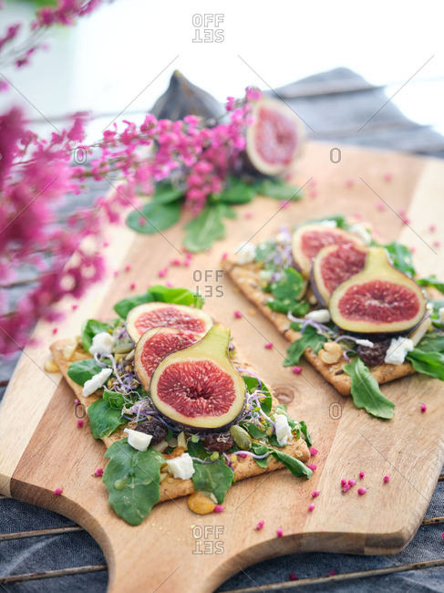 Homemade tasty colorful open sandwiches with slices of fig and pieces of cheese on crisp rye bread among aromatic green leaves of rocket salad and pink flowers