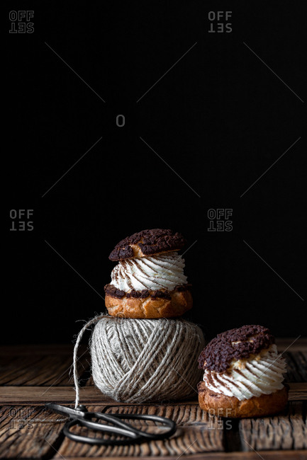 Homemade choux pastries with cream and chocolate scissors and ball of yarn arranged on texture wooden surface against black background