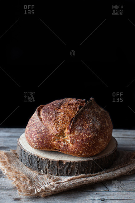 Loaf of fresh country sourdough bread placed on piece of wood on shabby table against black background