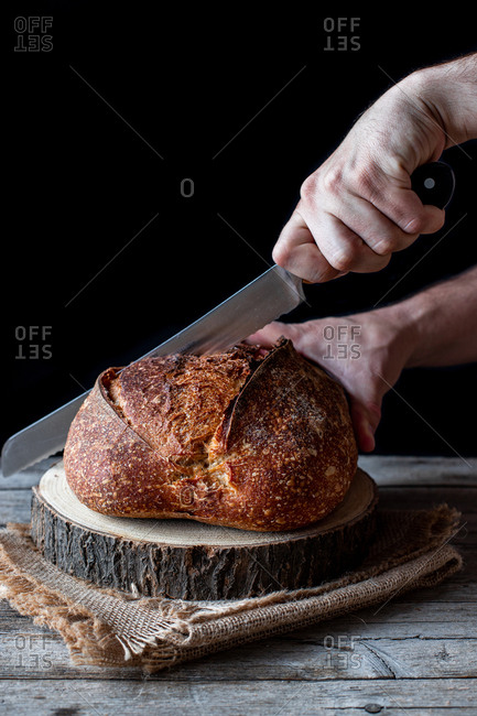 Unrecognizable person using knife to cut loaf of fresh sourdough bread on piece of wood against black background