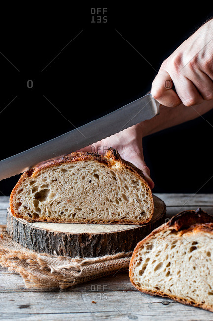 Unrecognizable person using knife to cut loaf of fresh sourdough bread on piece of wood against black background