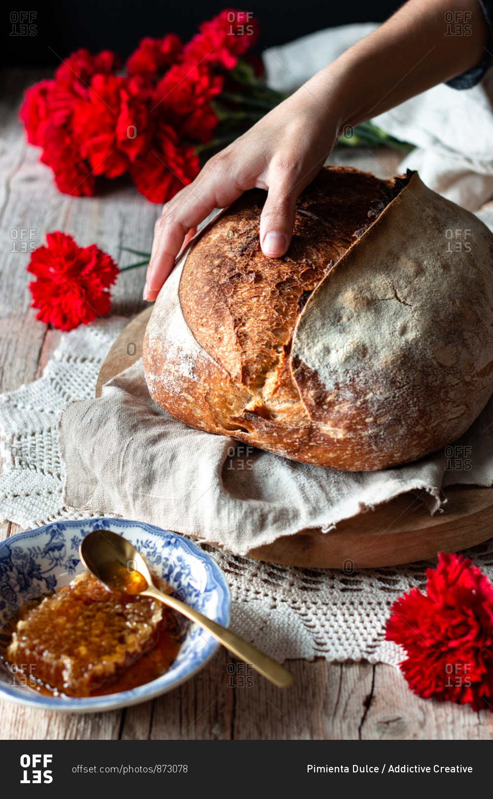 Unrecognizable person putting loaf of sourdough bread on rustic table near honeycomb and bouquet of red carnations