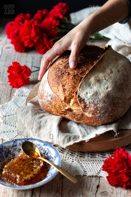 Unrecognizable person putting loaf of sourdough bread on rustic table near honeycomb and bouquet of red carnations