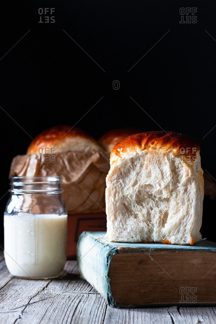 Small jar of fresh milk and buns of fresh bread placed on timber table near old recipe book against black background