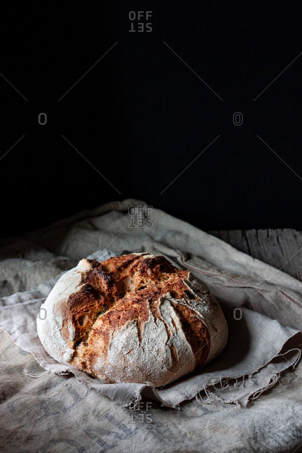 Loaf of fresh country sourdough bread placed on piece of wood on shabby table against black background