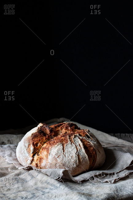 Loaf of fresh country sourdough bread placed on piece of wood on shabby table against black background