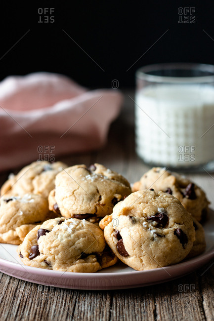 Plate of tasty vegan cookies with chocolate chips placed on lumber table near blurred milk and cloth against black background