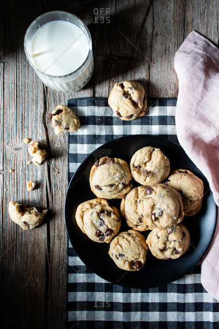 Plate of tasty vegan cookies with chocolate chips placed on lumber table near blurred milk and cloth against black background