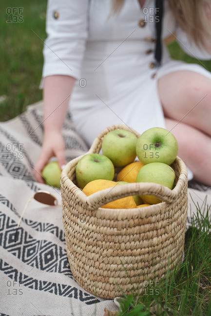 Beautiful woman in a white dress with a wicker basket of apples sits on a green grass on a plaid