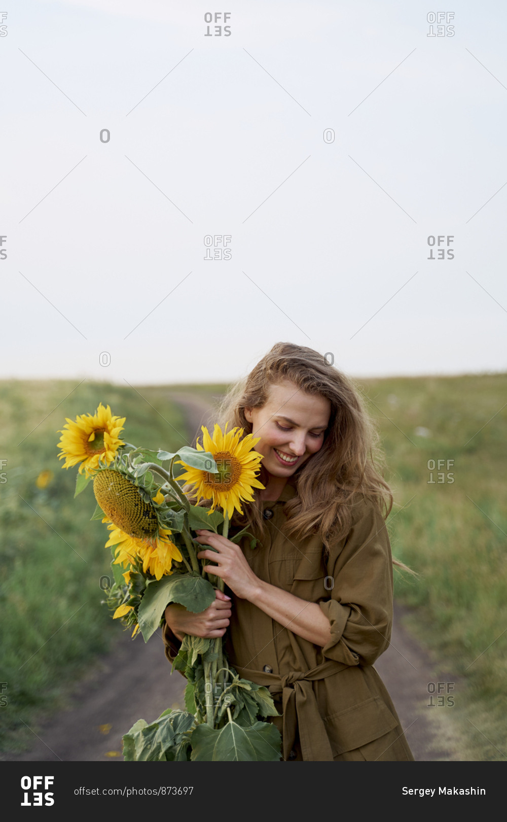 Beautiful woman traveler blogger in a jacket and yellow sunflowers on the road in a wheat field