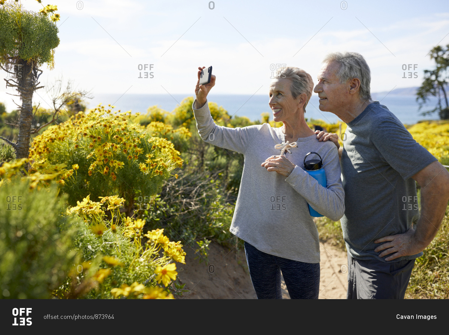 Smiling senior couple taking selfie through smart phone while standing on landscape by sea against sky during sunny day