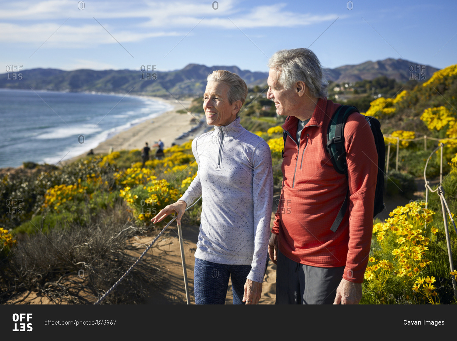 Smiling senior couple talking while walking on mountain by beach against sky during summer