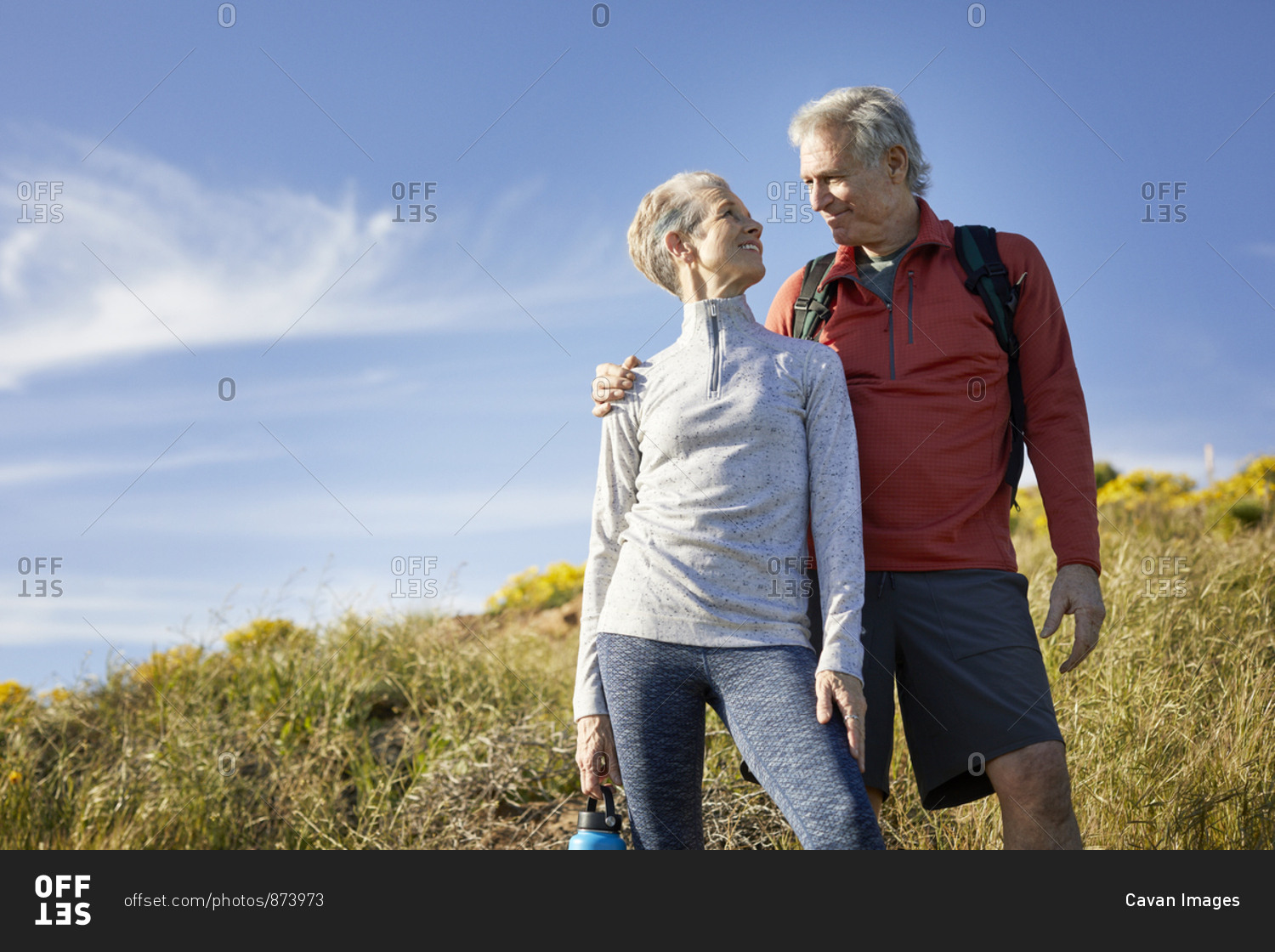 Low angle view of smiling senior couple looking at each other while standing on cliff against blue sky during sunny day