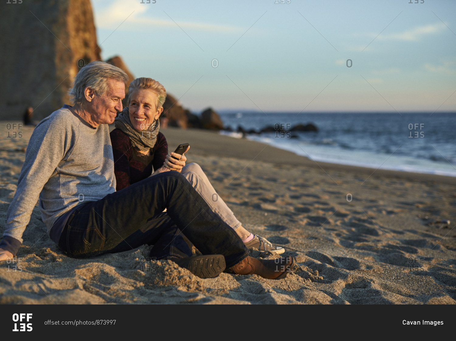 Senior couple using phone while sitting on sand at beach during sunset