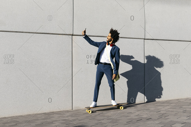 Young businessman riding skateboard along a wall taking a selfie