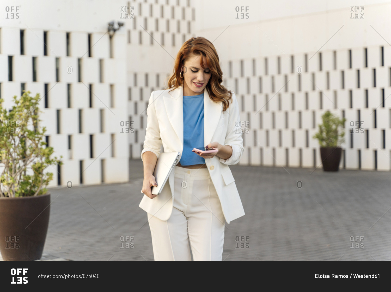 Businesswoman in white pant suit- carrying laptop- using smartphone