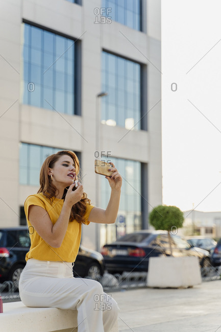 Businesswoman sitting in the city- using smartphone to put on lipstick