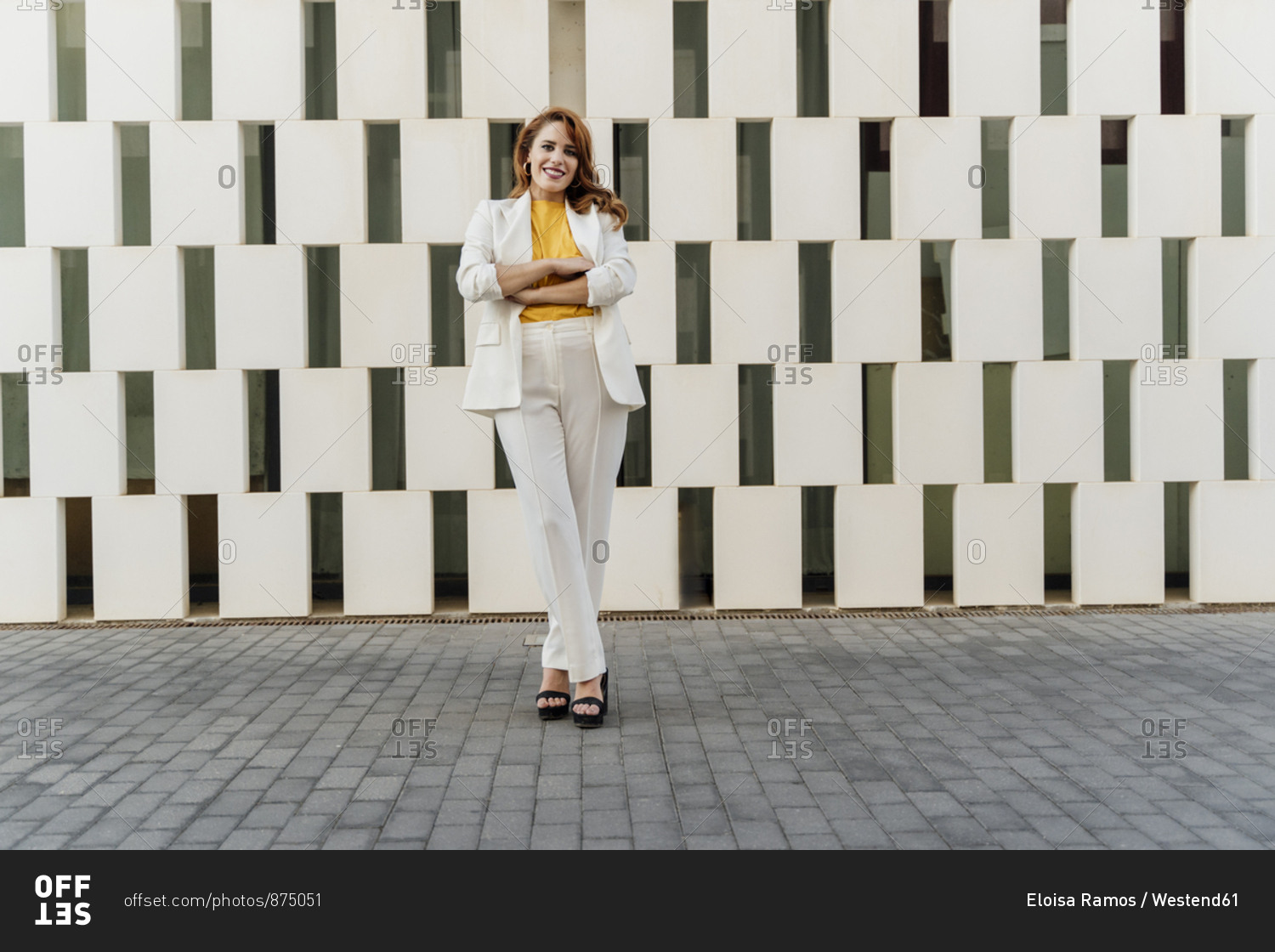 Businesswoman in white pant suit- standing in front of facade- with arms crossed