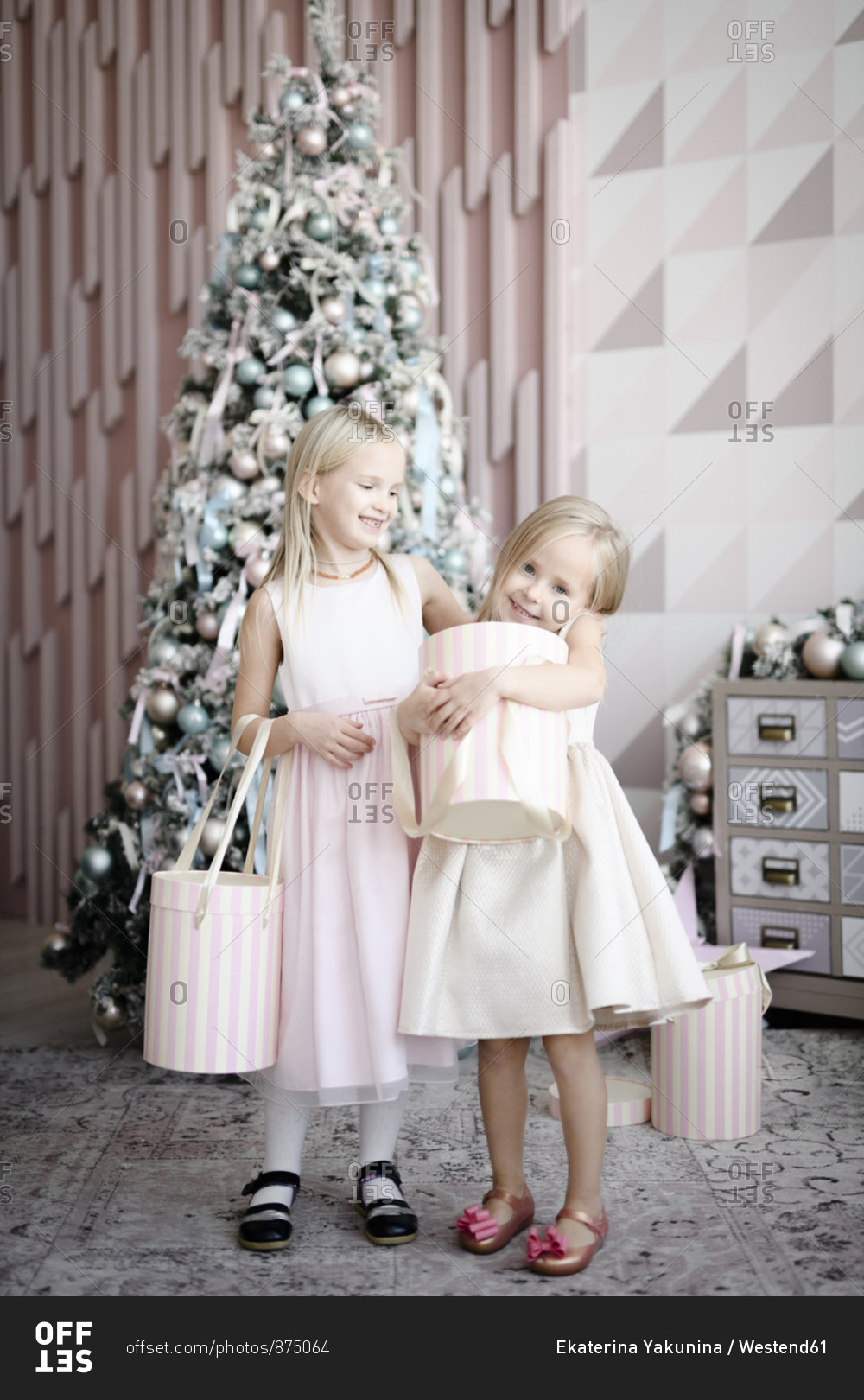 Portrait of two happy little girls standing in front of lighted Christmas tree with gift boxes