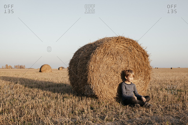 Boy sitting on stubble field leaning against hay bale