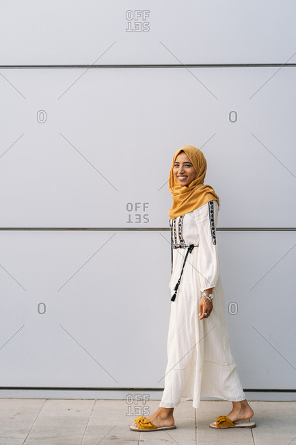 Young Muslim woman wearing yellow hijab and walking in front of a white wall