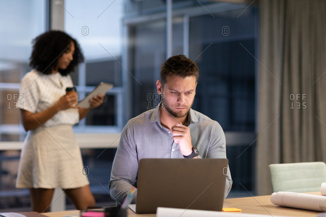 Young Caucasian professional man working after hours in a modern office at a desk using a laptop computer and mixed race female coworker standing in the background using a tablet