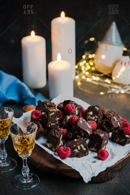 Homemade dark chocolate fudge with fruits and nuts on wooden board over dark background, candles and festive decorations