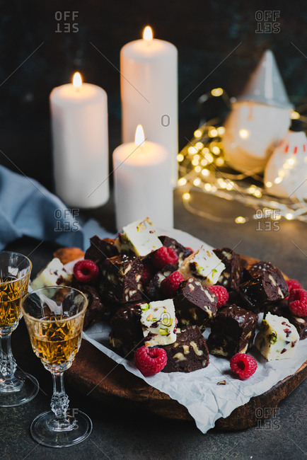 Homemade white and dark chocolate fudge with fruits and nuts on wooden board over dark background, candles and festive decorations