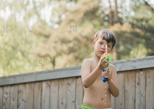 Portrait of boy spraying water from squirt gun while standing against wooden fence at yard