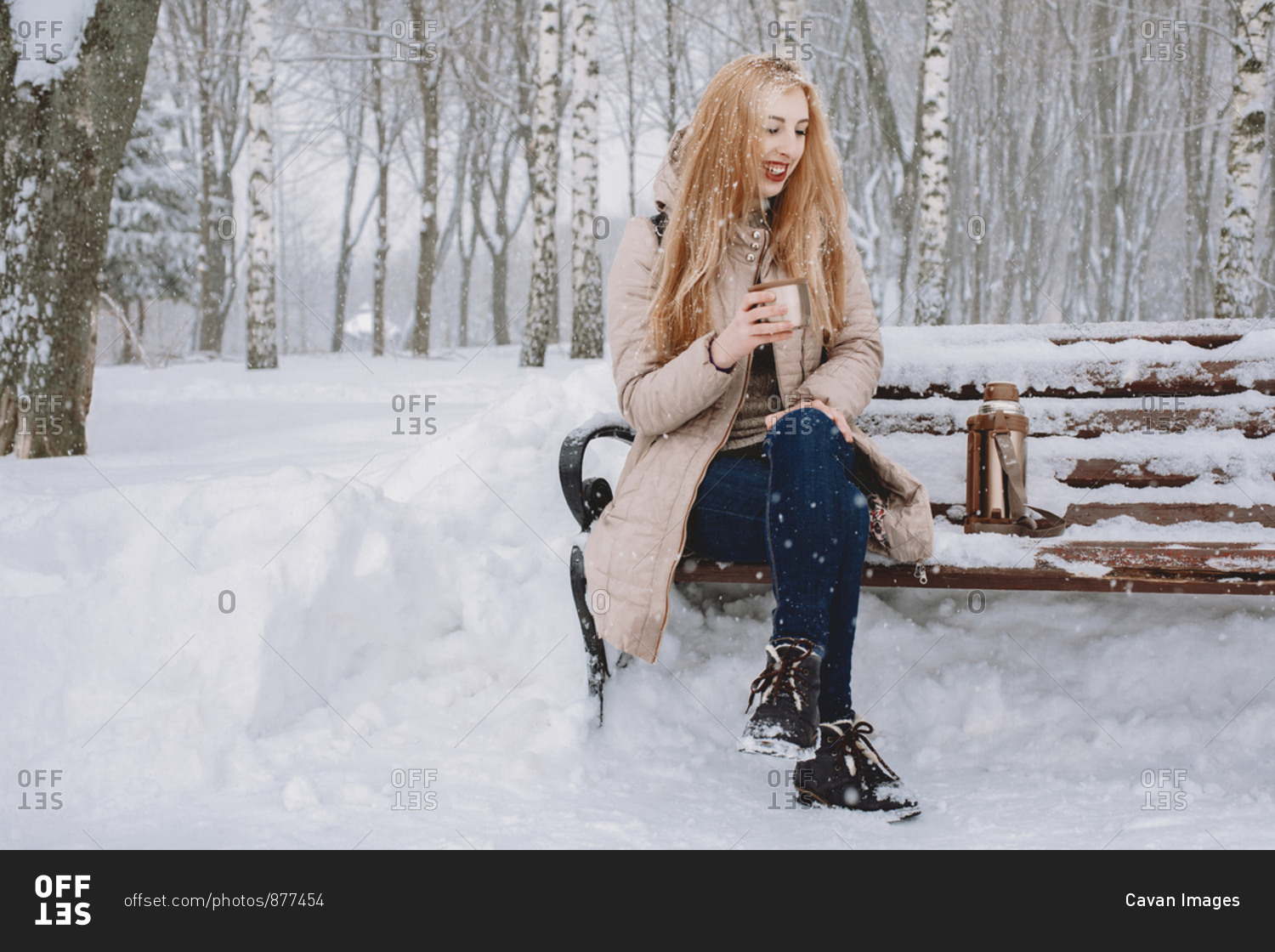 Happy woman having tea while sitting on bench against trees in park during winter