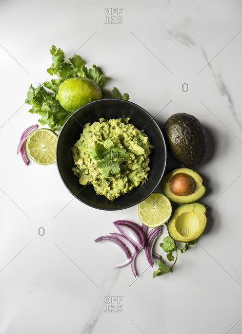 Bowl of guacamole surrounded by it's ingredients on a marble counter.