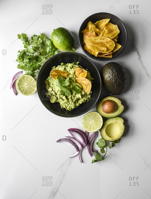 Bowl of guacamole and chips with it's ingredients on a marble counter.