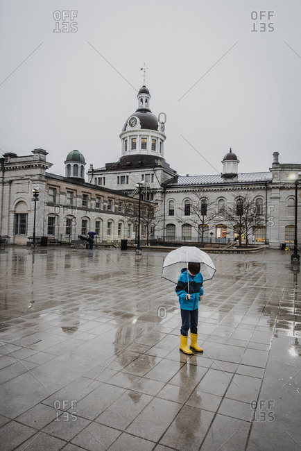 Boy in yellow rain boots holding umbrella in the rain in city square.