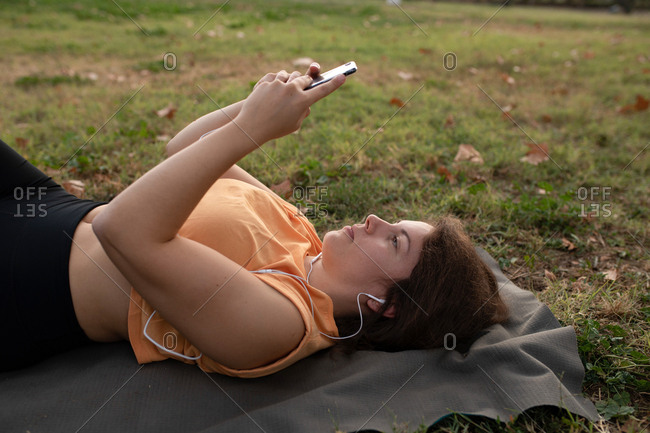 Young woman lying on the ground resting after doing a yoga, and listening to music from her phone