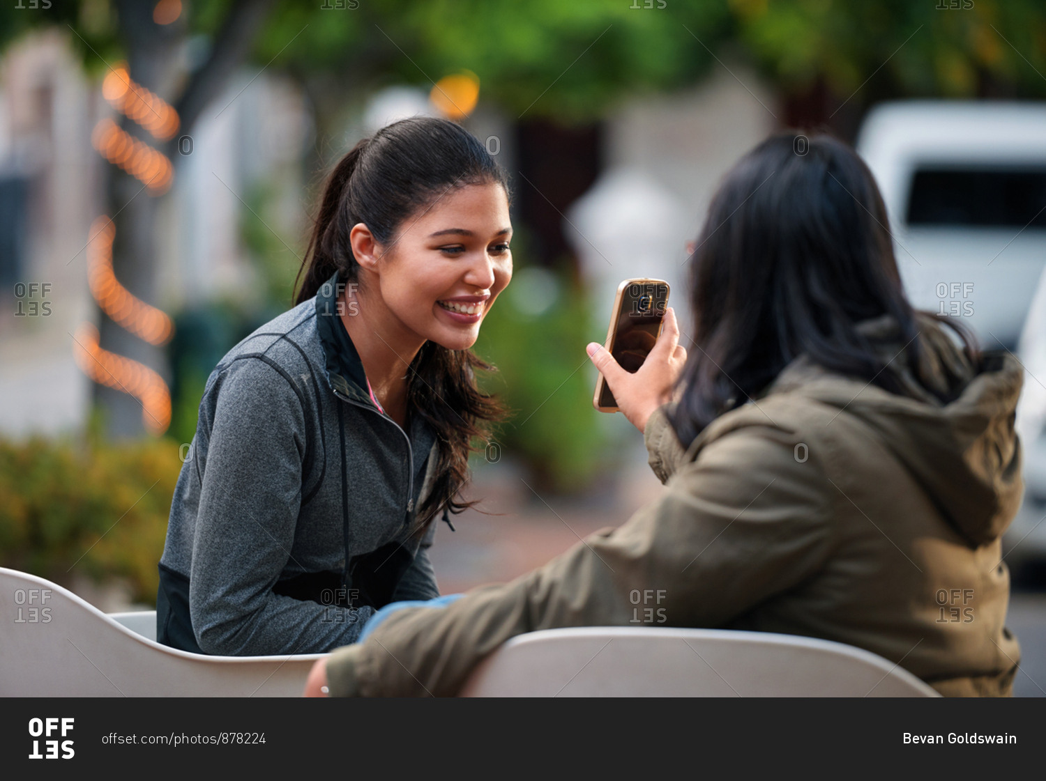 Woman showing smartphone to best friend smiling happy looking at mobile phone best friends sitting on bench in city
