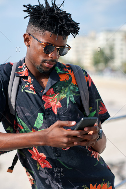 African american travel man using smartphone on beach texting with mobile phone sharing summer vacation wearing colorful Hawaiian shirt