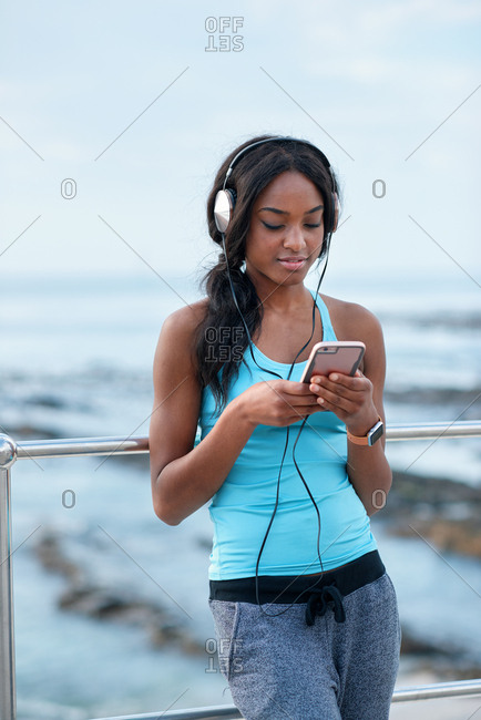 Young african american woman using smartphone on beach female jogger listening to music wearing headphones relaxing by the sea