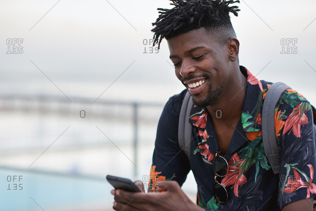 African american travel man using smartphone on beach texting with mobile phone sharing summer vacation wearing colorful Hawaiian shirt