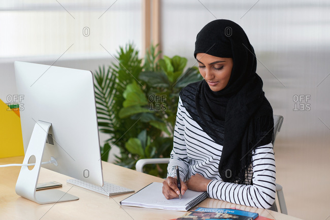 Muslim business woman writing ideas in notebook journal wearing hijab headscarf in office