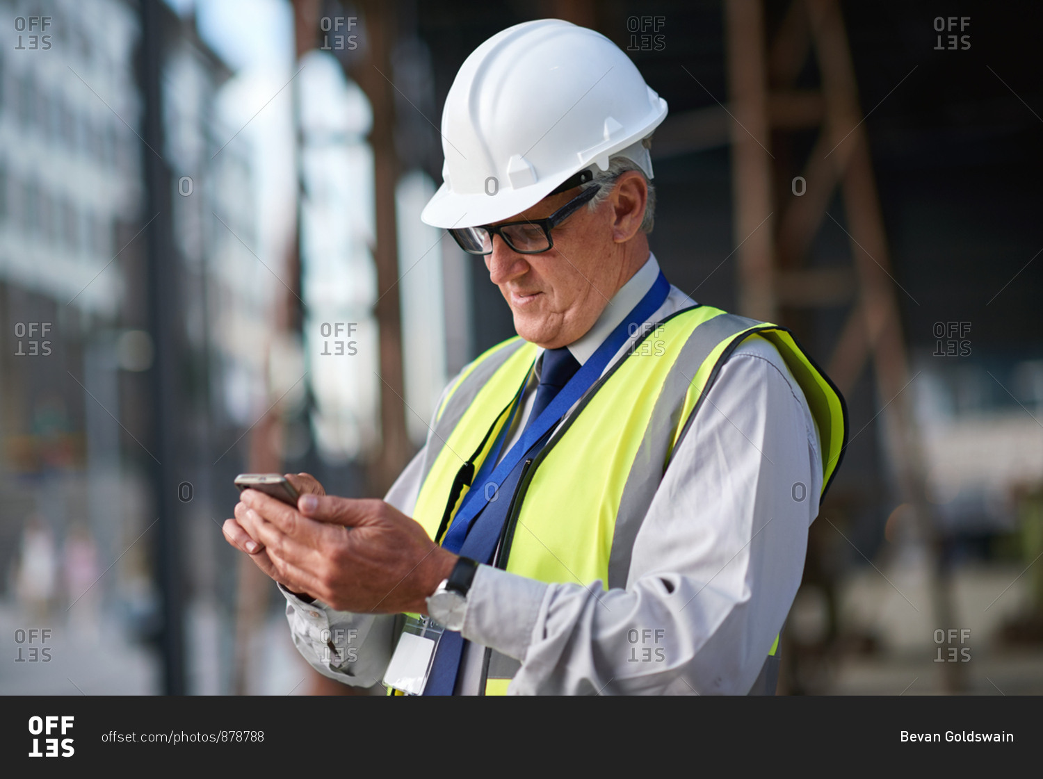 Mature construction worker man using smartphone browsing messages on site wearing hard hat and reflective vest in city